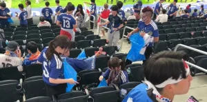 Fans in blue jerseys sit and stand in stadium seats, some cleaning up with blue bags after a game as others chat and walk by on the field side