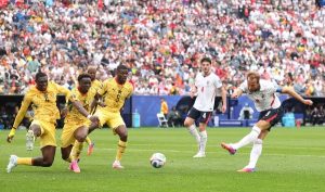 Soccer players in yellow and white kits sprint toward a ball on a green pitch as a crowded stadium watches from the stands.