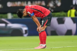 Soccer player in red kit bending over with hands on knees on a green pitch, wearing a blue captain's armband and pink shoes.