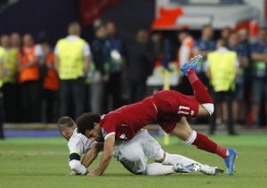 Two soccer players fall over during a tackle, red jersey over white, on a green pitch with a blurred crowd in the background