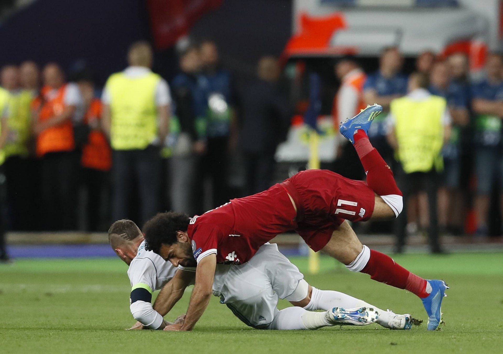Two soccer players fall over during a tackle, red jersey over white, on a green pitch with a blurred crowd in the background