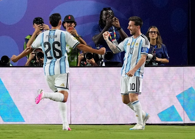 Argentina players celebrate on the field after a goal, with photographers watching in the background.
