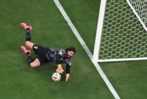 Soccer goalkeeper in a black kit dives to stop the ball near the goal line, glove reaching for it.