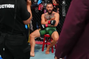 6c4c9b10-7dad-11f1-9eff-039a8b591bbf - 實發體育 MMA fighter with tattoos sits shirtless on a red stool inside the cage, wearing green shorts and UFC gloves during a break.