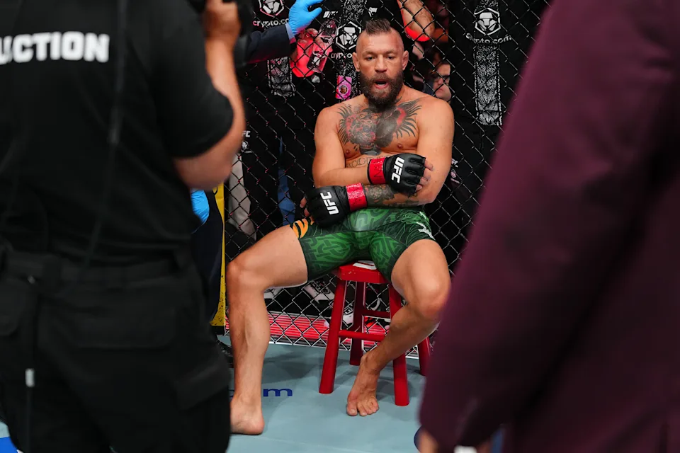 MMA fighter with tattoos sits shirtless on a red stool inside the cage, wearing green shorts and UFC gloves during a break.