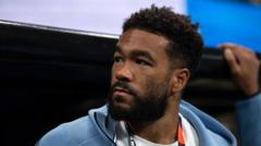 cover-8460-1783209662jpg - 實發體育 Man with short curly hair and beard wearing a light blue jacket sits courtside, looking off to the side.