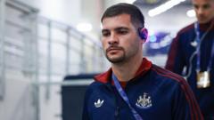 Man in a dark blue sports jacket with a crest, wearing a red lanyard, walking down a hallway in a building.