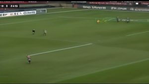 Soccer field during a match: a player in red and white near the foreground, two players running toward the center, and the goalie near the goal in the background with advertising boards around.
