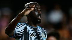 Close-up of a Black soccer player in a blue-and-white striped jersey, raising his right hand to shield his eyes during a match.
