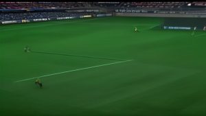 cover-9184-1786323626jpg - 實發體育 Soccer match on a green field with three players visible: one near the bottom-left, two toward the center-right, with empty stadium seats in the background.