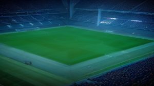 Empty football stadium at night with a bright green pitch and surrounding seating.