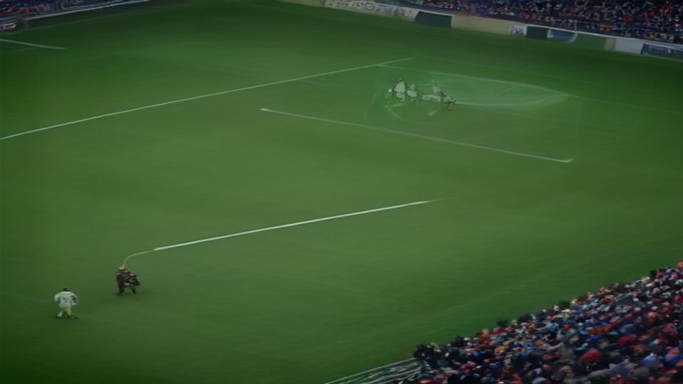 cover-9230-1786669231jpg - 實發體育 Aerial view of a soccer stadium with players walking across the green field toward the left while spectators fill the stands behind them, near a goal area in the distance.