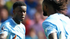 Two soccer players in light blue jerseys stand face-to-face on the field during a match, with a blurred crowd in the background.