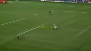 Soccer match: a white-jersey player advances with the ball on a green field, a yellow curved trail marks the ball's path.