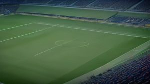 cover-9415-1788397229jpg - 實發體育 Empty football stadium with a green pitch and surrounding blue seating, no players visible.