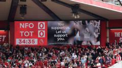 Crowd of football supporters in a red stadium, with large red banners and an LED screen above the stands displaying a message.